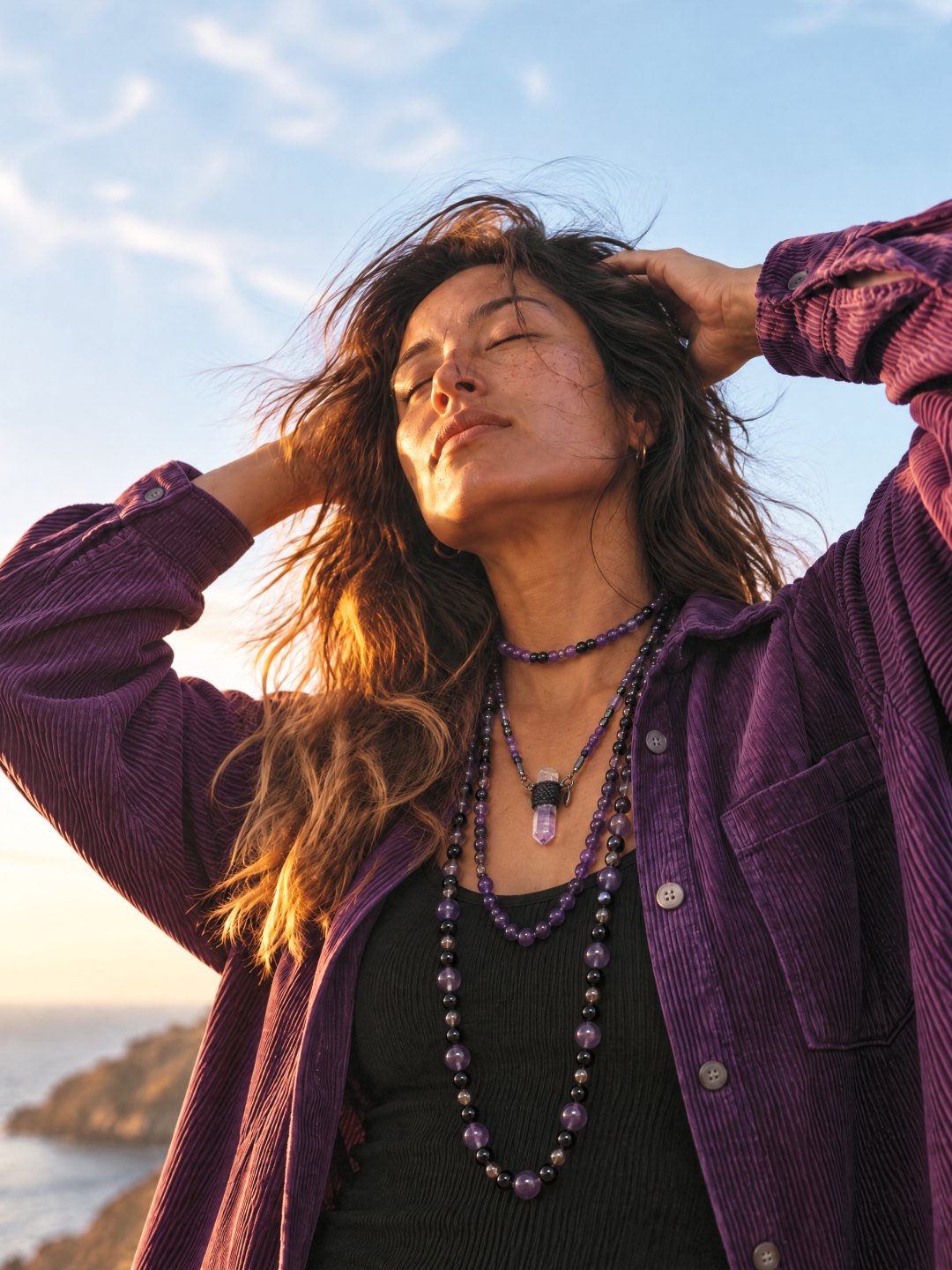 A woman on the coast with her arms raised, wearing layered Clink necklaces