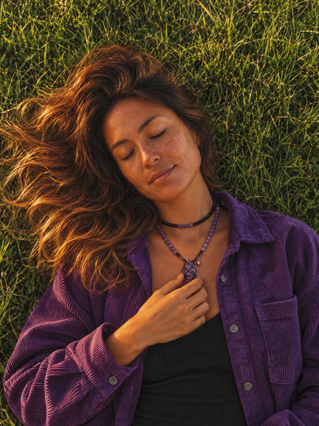 A woman lying in the grass, eyes closed, her hand on a netted Clink amethyst pendant